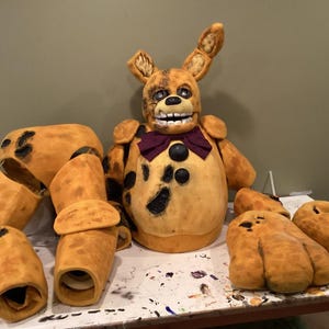 Puede incluir: Un disfraz de Springtrap amarillo y negro, con una cabeza grande con orejas de conejo, una pajarita morada y botones negros. El disfraz incluye piezas para brazos y piernas, y cubiertas para los pies grandes. El disfraz es de espuma.