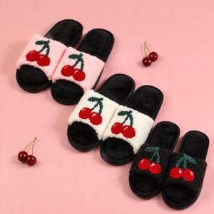 May include: Three pairs of open-toe slippers featuring a fuzzy texture and a cherry graphic on the strap. The slippers come in light pink, white, and black, all with black footbeds. Two loose cherries are placed near the slippers.