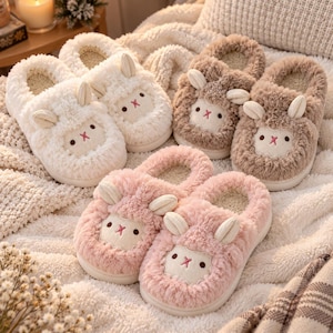 May include: Three pairs of plush slippers in white, brown, and pink. Each slipper features a fuzzy texture, two small upright ears, and a stitched face with two dark eyes and a pink cross-shaped nose. The interior is lined with a soft material.