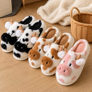 May include: Five plush slippers shaped like cows, arranged in a row. The slippers feature soft white faux fur with patches in black, brown, and pink. Each slipper has a fuzzy snout, small ears, and two tiny horns, with matching colored inner linings.