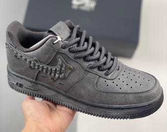 May include: Gray suede Air Force 1 sneakers with decorative metal accents. The sneakers feature a dark gray sole, laces, and a textured Swoosh logo. The shoe has a perforated toe box and is displayed with a dark gray box in the background.