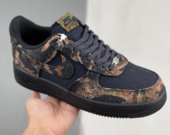 May include: Black and camouflage Air Force 1 sneakers. The shoe features a black sole, laces, and heel. The upper is a combination of black and a camouflage pattern in shades of brown, tan, and green. The Nike swoosh is visible on the side.
