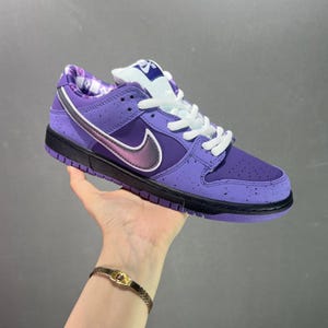 Puede incluir: Zapatilla deportiva morada y negra con cordones blancos y un logotipo swoosh morado y negro. La zapatilla tiene un diseño moteado en la parte superior y una suela negra. La lengüeta presenta un diseño blanco y morado.