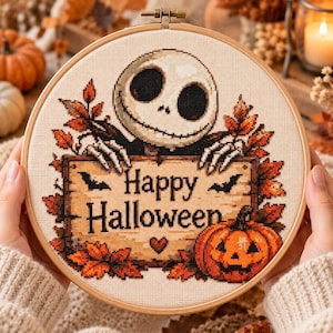 Puede incluir: Un bastidor de bordado circular de madera que contiene un diseño de punto de cruz de un personaje esquelético sosteniendo un letrero de madera que dice Happy Halloween. El diseño incluye hojas de otoño, dos murciélagos negros y una calabaza.