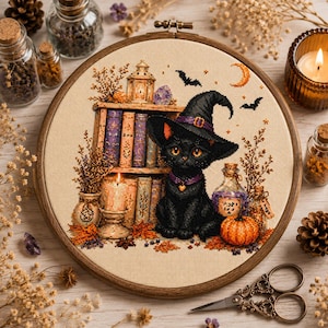 May include: A circular wooden embroidery hoop containing a cross-stitch design of a black cat wearing a purple-banded witch hat. The cat sits before a bookshelf with candles, a pumpkin, a potion bottle, and bats against a cream-coloured fabric.