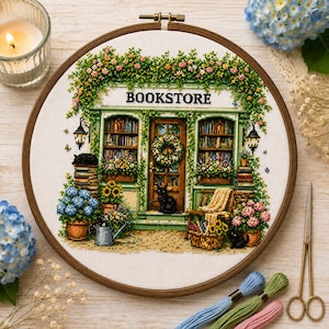 Könnte beinhalten: Ein runder Stickrahmen mit einem Kreuzstichmuster einer Buchhandlung. Das Design zeigt ein grünes Gebäude mit dem Wort BOOKSTORE, Bücherregale, einen Kranz an der Tür, Topfblumen und drei schwarze Katzen auf dem gestickten Bild.