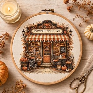 Puede incluir: Un bastidor de bordado circular de madera que contiene un patrón de punto de cruz de una tienda con la etiqueta PUMPKIN SPICE CAFE. El diseño presenta un toldo a rayas naranjas y blancas, follaje otoñal, calabazas y asientos exteriores.