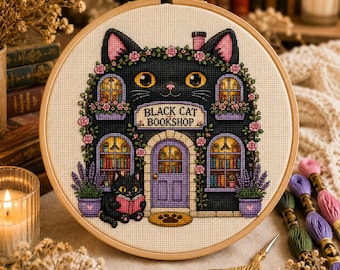 May include: A cross-stitch embroidery in a wooden hoop featuring a black cat shaped building with the text BLACK CAT BOOKSHOP. The design includes a purple door, arched windows with books, pink flowers, lavender plants, and a small cat reading.