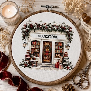 Könnte beinhalten: Eine Kreuzstichstickerei in einem runden Holzrahmen, die eine Buchhandlung mit dem Schriftzug BOOKSTORE über der Tür zeigt. Das Motiv umfasst schneebedecktes Grün, rote Beeren, Bücherstapel und einen Kranz an der Eingangstür.
