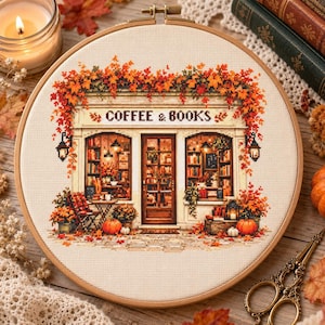 May include: A circular wooden embroidery hoop containing a cross-stitch pattern of a storefront labelled COFFEE & BOOKS. The shop is decorated with autumn foliage, pumpkins, and warm lighting, featuring shelves of books visible through the windows.