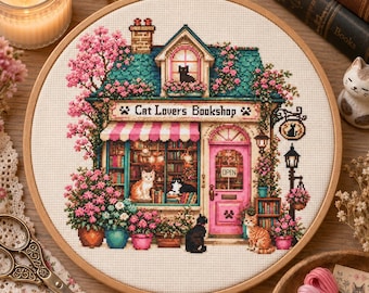 May include: A circular embroidery hoop containing a cross-stitch pattern of a bookstore titled Cat Lovers Bookshop. The design features a building with a teal roof, pink door, and multiple cats, surrounded by pink flowering bushes and potted plants.