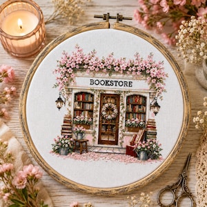 Puede incluir: Un bastidor de bordado circular de madera que contiene un diseño de punto de cruz de la fachada de una librería. La tienda cuenta con estanterías llenas de libros, una puerta central con una corona y enredaderas de flores rosas sobre la parte superior. La palabra BOOKSTORE está bordada en letras negras sobre la entrada.