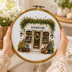 Könnte beinhalten: Ein runder Stickrahmen aus Holz mit einem Kreuzstichmotiv einer Buchhandlung. Das Geschäft zeigt eine weiße Fassade, mit Büchern gefüllte Regale, eine mittige Tür mit einem Kranz, hängende Laternen und rankende Blumenpflanzen.