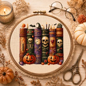 May include: A cross-stitch embroidery in a round wooden hoop featuring a row of six colorful, detailed books with Halloween-themed illustrations including skulls, pumpkins, a witch hat, a clock, and a heart, all set against a light fabric background.