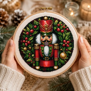May include: A circular embroidery hoop containing a cross-stitch pattern of a nutcracker soldier centered within a wreath of green holly leaves, red berries, and gold ornaments. The nutcracker wears a red and green uniform with gold accents.