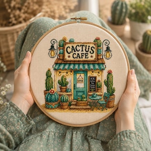 May include: A circular embroidery hoop containing a cross-stitch design of a storefront labelled CACTUS CAFE. The scene features a teal door, a striped awning, various potted cacti, two hanging lanterns, and a small table with a chair.