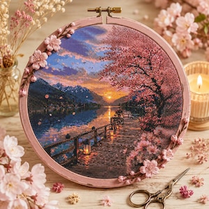 May include: A circular embroidery hoop frame in light pink, decorated with small pink fabric flowers. The fabric inside displays a cross-stitch landscape of a sunset over a lake, a wooden pier with glowing lanterns, and a blooming cherry tree.