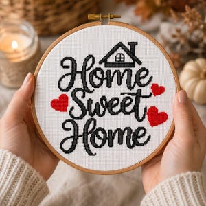 Op de afbeelding: Een ronde houten borduurring met witte stof waarop de tekst Home Sweet Home in zwarte kruissteek is geborduurd. Het ontwerp bevat een klein zwart huisicoon boven de tekst en drie rode, in kruissteek geborduurde hartjes rond de woorden.