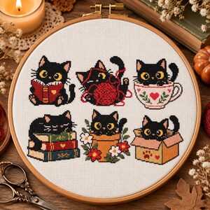 May include: A circular wooden embroidery hoop containing a white fabric panel with six cross-stitched black cats. The cats are depicted in various activities: reading a book, playing with red yarn, sitting in a teacup, resting on books, sitting in a flower pot, and hiding in a cardboard box.