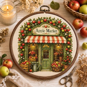 May include: A circular embroidery hoop containing a cross-stitch design of a storefront labelled Apple Market. The shop features a red and white striped awning, green trim, and a display of apples in crates and baskets, surrounded by apple tree branches.