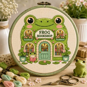 Può includere: Un telaio da ricamo circolare contenente un disegno a punto croce di un edificio a forma di rana con la scritta FROG BOOKSHOP. Il design presenta una porta centrale, finestre ad arco con librerie e una rana che legge un libro.