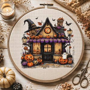 May include: A circular wooden embroidery hoop containing a cross-stitch pattern of a Halloween-themed house. The house features a witch hat roof, purple striped awnings, glowing windows, jack-o'-lanterns, a small ghost, and flying bats.