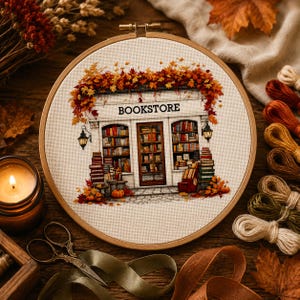 May include: A circular wooden embroidery hoop containing a cross-stitch design of a bookstore storefront. The shop features shelves filled with books, a central door, and a sign reading BOOKSTORE, all framed by vibrant orange and red autumn leaves.