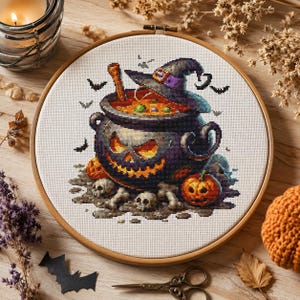May include: A circular wooden embroidery hoop containing a cross-stitch pattern of a dark purple cauldron with a glowing orange jack-o'-lantern face. The cauldron features a witch's hat, small skulls, bones, and pumpkins, surrounded by tiny black bats.