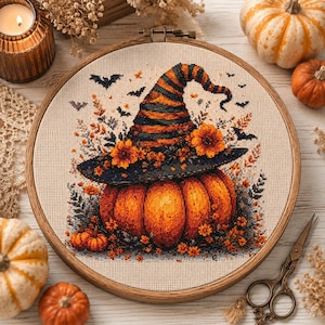Può includere: Un telaio da ricamo circolare in legno contenente un disegno a punto croce di una grande zucca arancione che indossa un cappello da strega a righe. Il cappello è decorato con fiori arancioni e la scena è circondata da piccoli pipistrelli.