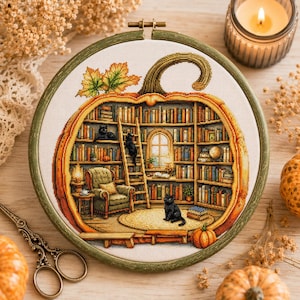 May include: A cross-stitch embroidery in a circular wooden hoop featuring a large orange pumpkin outline filled with a detailed library scene. The interior includes bookshelves, a green armchair, a ladder, and three black cats on a rug.