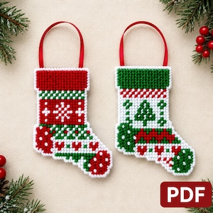Peut inclure: Deux décorations de Noël en forme de chaussettes brodées au point de croix avec des rubans rouges. L'une est principalement rouge avec des motifs verts et blancs, l'autre est blanche avec des motifs verts et rouges, incluant un sapin.