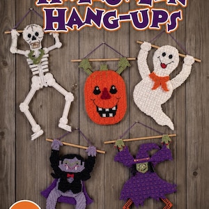 May include: Five crocheted Halloween wall hangings featuring a white skeleton, an orange pumpkin, a white ghost, a vampire in a purple cape, and a witch in a purple dress, all suspended from wooden dowels with the text Halloween Hang-Ups.