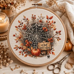 May include: A circular wooden embroidery hoop containing a cross-stitch design on cream fabric. The pattern features a central spider web, orange pumpkins, autumn leaves, red berries, and a wooden sign with the text HOME SWEET HOME.