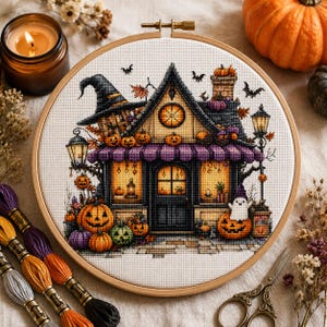 May include: A circular wooden embroidery hoop containing a cross-stitch pattern of a Halloween-themed house. The house features a witch hat roof, purple striped awning, glowing windows, pumpkins, and a small ghost. Embroidery floss and scissors are nearby.