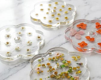Puede incluir: Cuatro posavasos transparentes en forma de flor con flores secas incrustadas en resina. Cada posavasos presenta una disposición diferente de flores en tonos blancos, amarillos, naranjas y verdes. Sobre una superficie de mármol blanco.