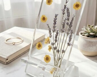 Puede incluir: Soporte transparente para teléfono de acrílico con lavanda seca y flores amarillas incrustadas. El soporte tiene una repisa incorporada para sostener un teléfono y se muestra sobre una superficie de mármol blanco. Una pulsera dorada y una suculenta están en el fondo.