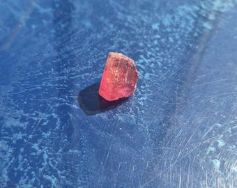 Puede incluir: Una pequeña piedra preciosa rosa, translúcida y en bruto, con una forma irregular y angular de bordes facetados. La piedra muestra un tono magenta vibrante que capta la luz, proyectando una sombra oscura sobre la superficie.