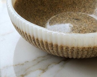 Puede incluir: Un cuenco decorativo con un interior texturizado similar a la arena. El borde es blanco con detalles acanalados verticales. El cuenco se encuentra sobre una superficie de mármol, reflejando la luz.