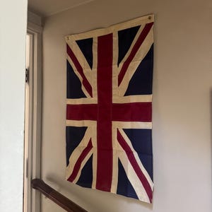 May include: A rectangular Union Jack flag featuring the traditional red, white, and blue color scheme. The flag is constructed with stitched fabric panels and includes two metal grommets at the top corners for mounting purposes.