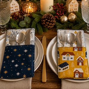 May include: Two rectangular fabric cutlery holders placed on white plates. The left holder is dark blue with gold stars. The right holder is mustard yellow with a white house pattern. Both contain a silver fork, spoon, and knife set.