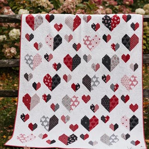 May include: A white quilt with a red border, featuring numerous heart shapes in red, black, and gray patterns. The hearts are arranged across the quilt's surface, creating a visually appealing design. The quilt is displayed outdoors.