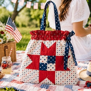 May include: A fabric tote bag featuring a red, white, and blue star quilt pattern on a white background with small stars. The bag has a ruffled top edge, dark blue handles with white hearts, and a matching blue ribbon tie on the side of the bag.