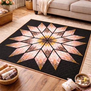 Pode incluir: Um edredom preto grande com um design central em forma de estrela. A estrela apresenta segmentos em forma de diamante em tons de rosa, pêssego e castanho, com detalhes dourados. O edredom está sobre um tapete claro.