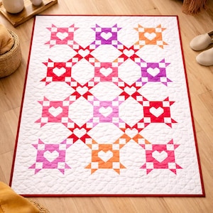 May include: A white quilt with a red border features a repeating pattern of hearts and star shapes in shades of red, pink, purple, and orange. The quilt is placed on a wooden floor.