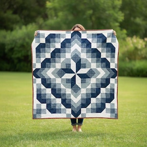 Puede incluir: Un edredón grande y cuadrado con un patrón geométrico en tonos azules y blancos. El edredón presenta un diseño central en forma de estrella y un borde con un ribete marrón. El edredón se sostiene sobre un fondo de hierba verde.
