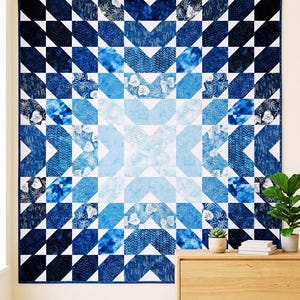 May include: A large, square quilt with a geometric pattern in shades of blue and white. The design features a central starburst motif surrounded by alternating blocks of dark and light blue triangles and squares. The quilt is displayed on a wall.