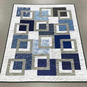 May include: A handmade quilt with a geometric design. The quilt features squares in shades of blue, gray, and white, arranged in a repeating pattern. The squares are framed by gray borders and set against a white background. The quilt is machine quilted with a wavy pattern.