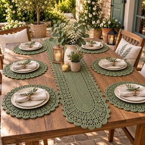 Puede incluir: Una mesa de comedor de madera con un camino de mesa de ganchillo verde salvia y cuatro manteles individuales redondos a juego. Cada cubierto incluye un plato de cerámica blanca, una servilleta de lino beige doblada y una ramita verde.