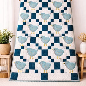 May include: A blue and white quilt with a repeating pattern of blue birds on a checkered background. The birds have yellow feet and beaks. The quilt is displayed on a wooden rack.