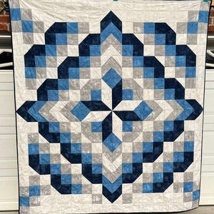 May include: A large, geometric quilt with a repeating pattern of squares and angled shapes in shades of blue, gray, and white. The quilt is displayed against a brick wall and has a black border.